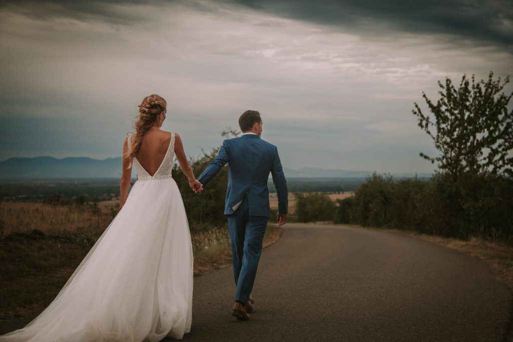 Un couple de mariés se tient la main. Ils viennent de se marier et avancent sur un chemin. On y voit la belle robe de mariée et le costume de marié.