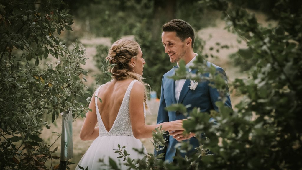 Un couple vient de se marier, ils se tiennent la main et  se sourient. Ils sont heureux. On y voit la robe de mariée, la coiffure, le costume de marié.