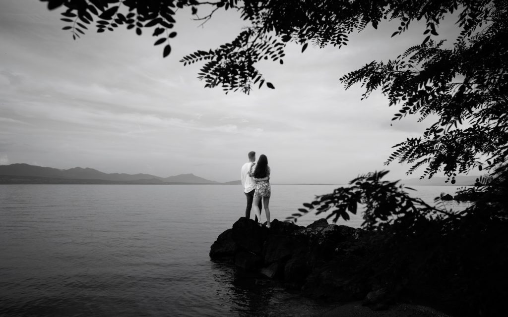 Un couple de futurs mariés lors d'une séance de fiançailles regarde le lac Léman et l'horizon, en Suisse. Ils sont sur des rochers. La photo est en noir et blanc.