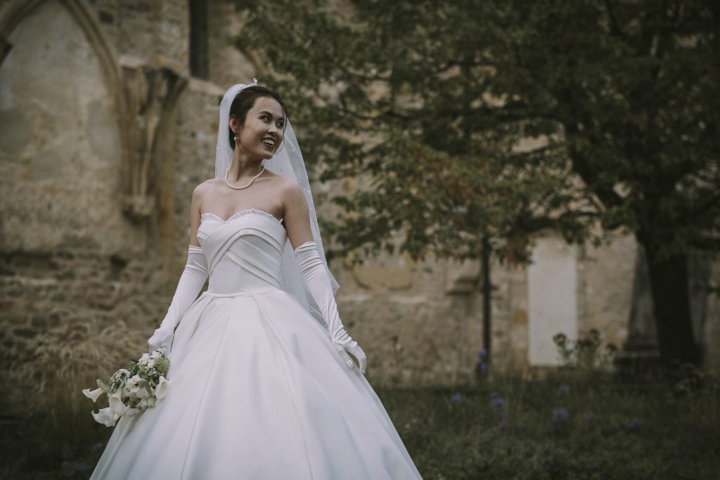 Une jeune femme vient de se marier. Elle est prise en photo devant l'abbatiale de Romainmôtier. On y voit sa robe de mariée ainsi que son bouquet, ses bijoux, ses gants blancs, son voile et son sourire.