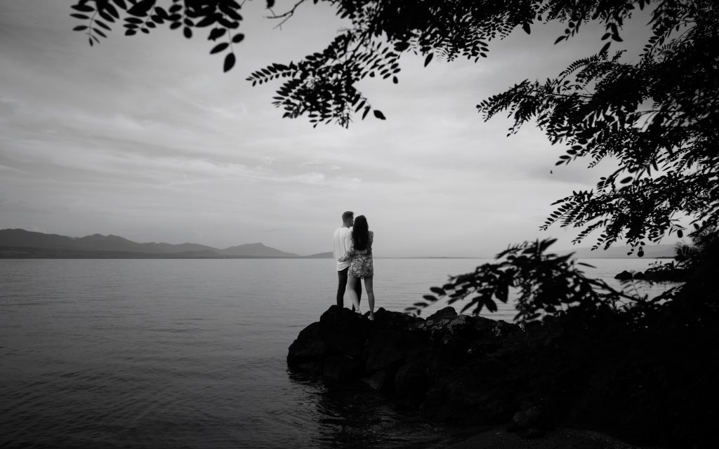 Un jeune couple de futurs mariés fait une séance de photos de couple. Ils sont face au lac Léman. On les voit de dos, enlacés. La photo est en noir et blanc.