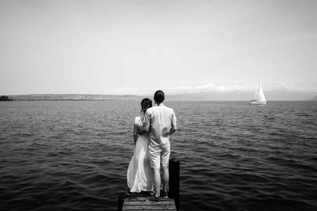 Deux jeunes mariés font face au lac de Neuchâtel. C'est une photo en noir et blanc devant le lac de Neuchâtel. On y voit la robe de mariée, ses bijoux, sa coiffure, le costume de marié.