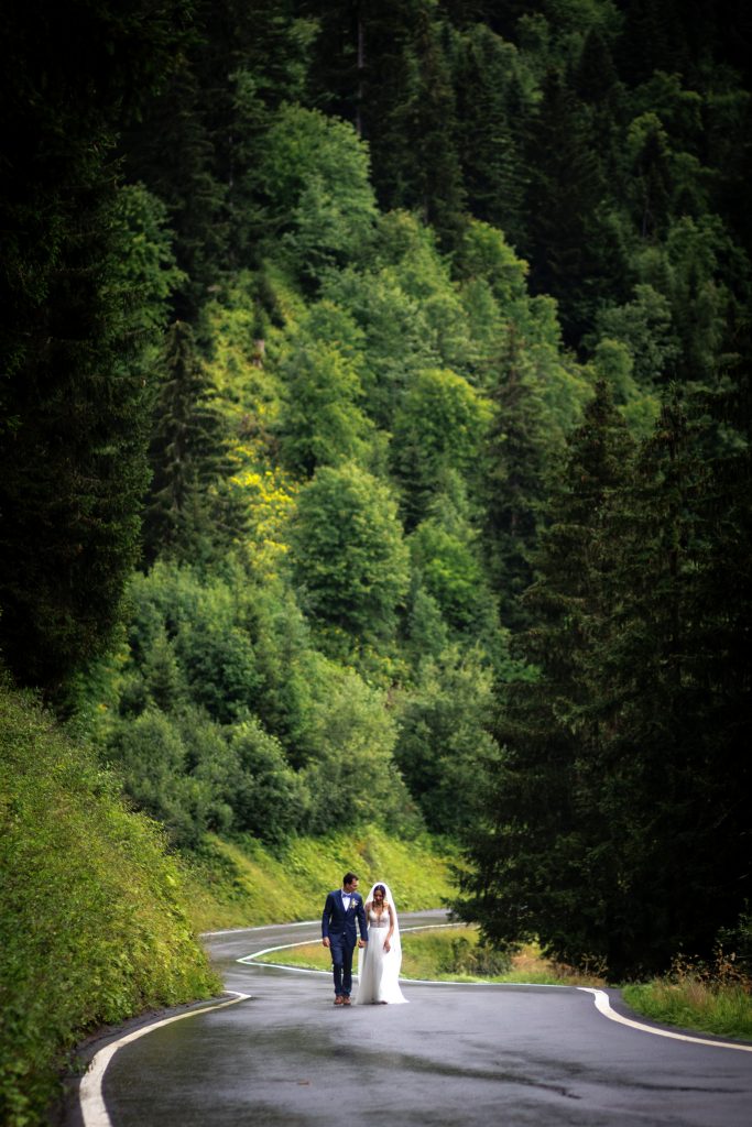 Un couple de jeunes mariés marche sur une route en plein milieu d'une forêt en montagne. C'est une photo en portrait, les arbres sont magnifiques. Ils se tiennent la main. Nous sommes à Champoussin.