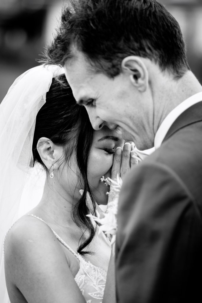 La mariée pleure d'émotions, le marié la tient par la taille, et sourit. Sur cette photo en noir et blanc, on voit la belle robe de mariée ainsi que ses bijoux et le costume du marié. Ils sont heureux. C'est une photo de couple.