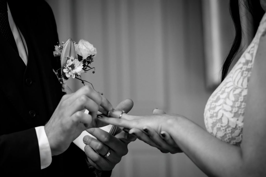 Le marié met la l'alliance au doigt de sa femme. Ils viennent de se marier à Lausanne. La photo est en noir et blanc. On voit la belle robe de mariée et le costume de marié.