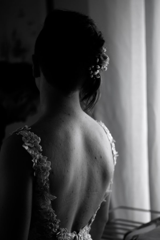 La future mariée vient d'enfiler sa belle robe de mariée, on la voit de dos, en noir et blanc, avec des fleurs dans les cheveux. 