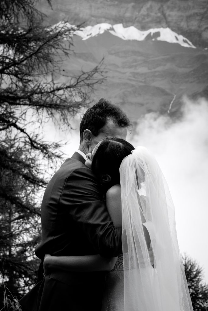C'est un mariage en Suisse, les mariés s'enlacent devant les montagnes enneigées. Sur cette photo en noir et blanc, on voit la belle robe de mariée, le costume de marié et les bijoux. 